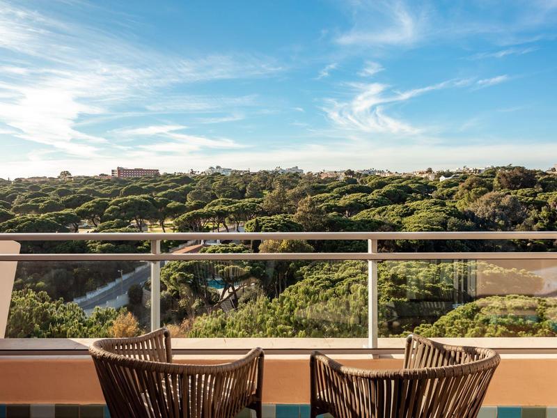 Balkon mit zwei Stühlen, Glasgeländer und grüner Baumlandschaft unter blauem Himmel.