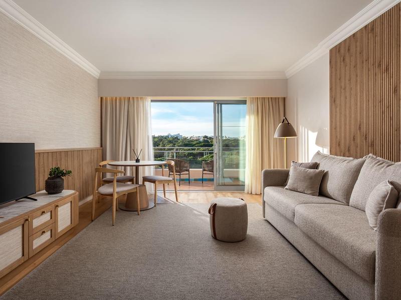 Helles Wohnzimmer mit grauem Sofa, Holzmöbeln, Teppichboden und Balkon mit grüner Aussicht.