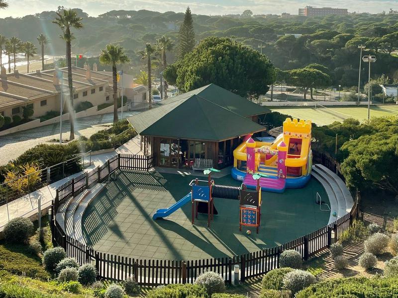 Spielplatz mit Klettergerüst und Rutsche, umgeben von Grün und Hügeln unter blauem Himmel.