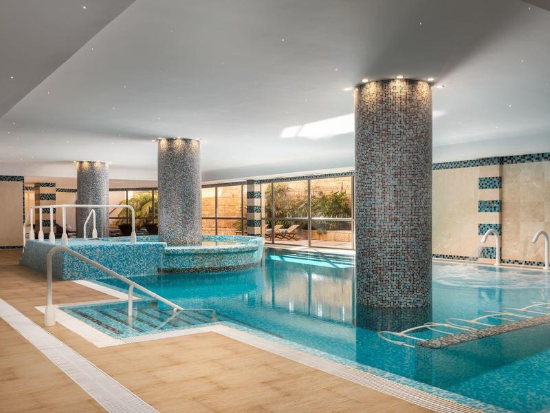 Hallenbad mit blauem Wasser, runden Säulen, Treppe und großen Fenstern, helle Decke und Wände.