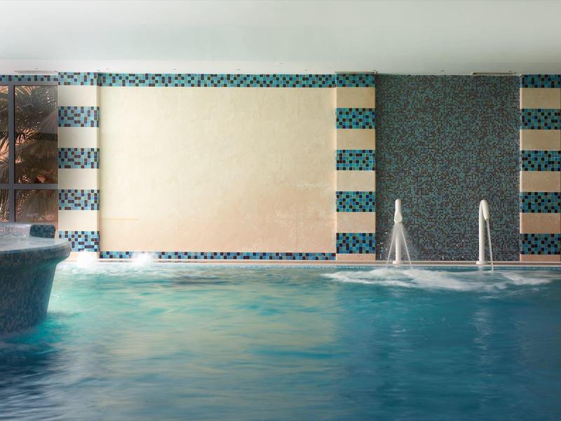 Innen-Schwimmbad mit klar blauem Wasser, Wasserfall und rutschigem Beckenrand.