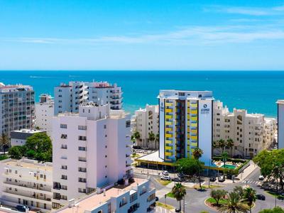 Helle Wohngebäude am Meer unter klarem blauem Himmel mit Straßen und Bäumen im Vordergrund.