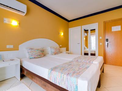 Hellgelbes Hotelzimmer mit Doppelbett, Nachttisch, Klimaanlage und Spiegel an der Tür.