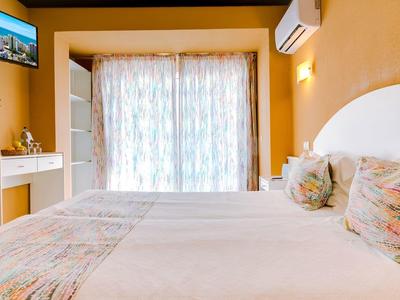 Helles Schlafzimmer mit großem Bett, bunten Kissen, TV an der Wand und Balkon mit Vorhängen.