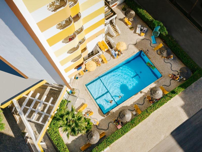 Überblick über einen rechteckigen blauen Pool, umgeben von Liegestühlen und Sonnenschirmen neben einem gelb-weißen Gebäude.