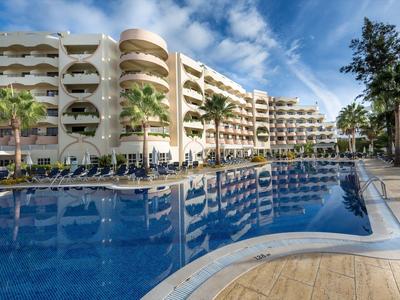 Modernes Hotel mit großem Pool und Liegestühlen unter blauem Himmel.