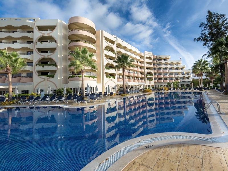 Nowoczesny hotel z basenem na zewnątrz i wieloma leżakami pod niebieskim niebem.