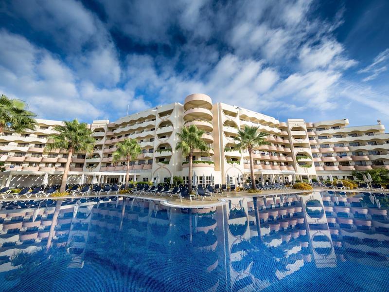 Nowoczesny hotel z basenem na zewnątrz i palmami pod niebieskim niebem z chmurami.