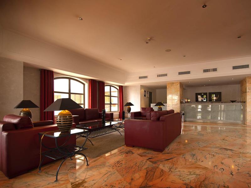 Duży salon hotelowy z kamienną podłogą, bordowymi sofami i dużymi oknami.