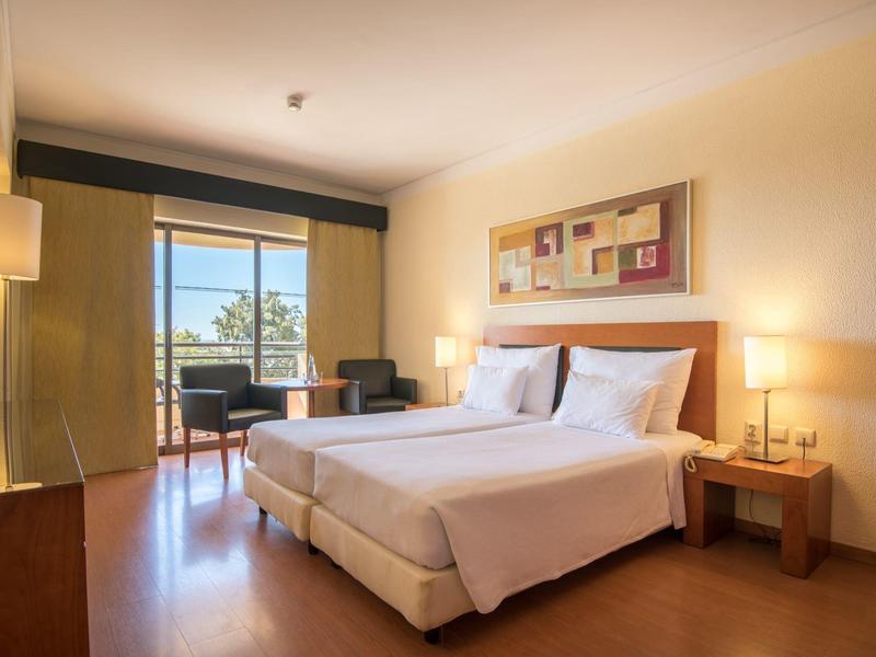 Jasny pokój hotelowy z podwójnym łóżkiem, biurkiem, lampą i balkonem z widokiem na zewnątrz.