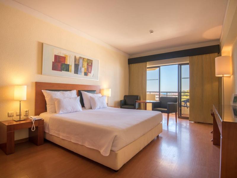 Jasny pokój hotelowy z dużym łóżkiem i balkonem z widokiem na morze.