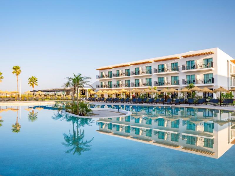 Modern hotel met groot zwembad, palmbomen en lichte blauwe lucht op de achtergrond.