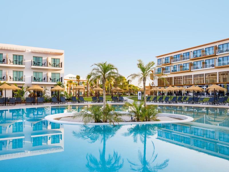 Modern hotelcomplex met groot zwembad en eiland met palmbomen onder heldere lucht.