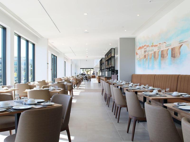 Licht, modern restaurant met rijen gedekte tafels en bruine stoelen bij grote ramen.