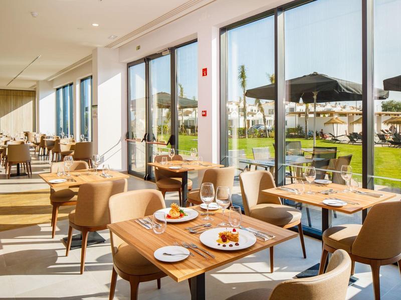 Lichte restaurantruimte met houten tafels en stoelen met uitzicht op een terras en tuin.