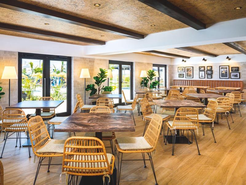 Licht, gezellig restaurant met houten tafels, rieten stoelen en grote ramen met uitzicht op planten.