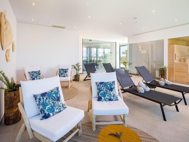 Moderne wellnessruimte met ligstoelen, zitmeubels en glazen sauna in lichte sfeer.