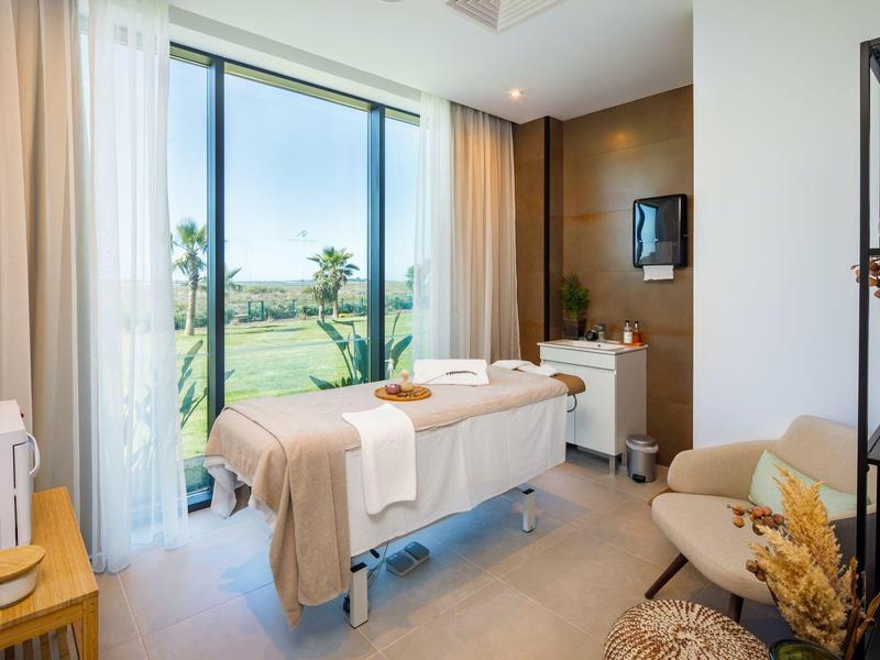 Lichte, modern ingerichte massageruimte met tafel voor groot raam met uitzicht op groen.