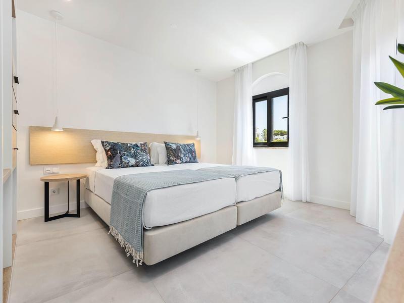 Nowoczesna, jasna sypialnia z łóżkiem podwójnym, stolikiem nocnym, dużym oknem i minimalistycznym wystrojem.