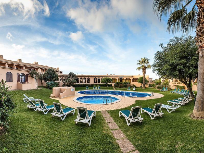 Hotel z okrągłym basenem, leżakami i ogrodem pod błękitnym niebem.