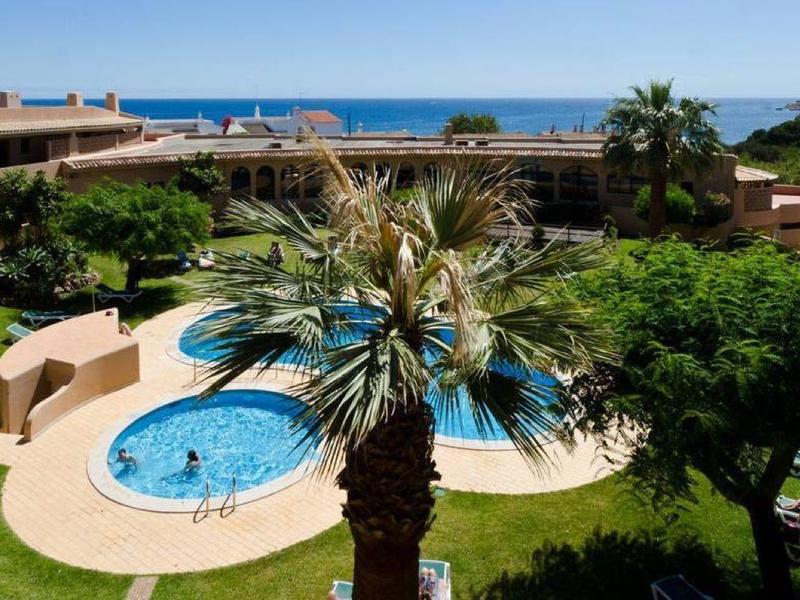 Kompleks hotelowy z okrągłym basenem, leżakami i widokiem na morze w tle.