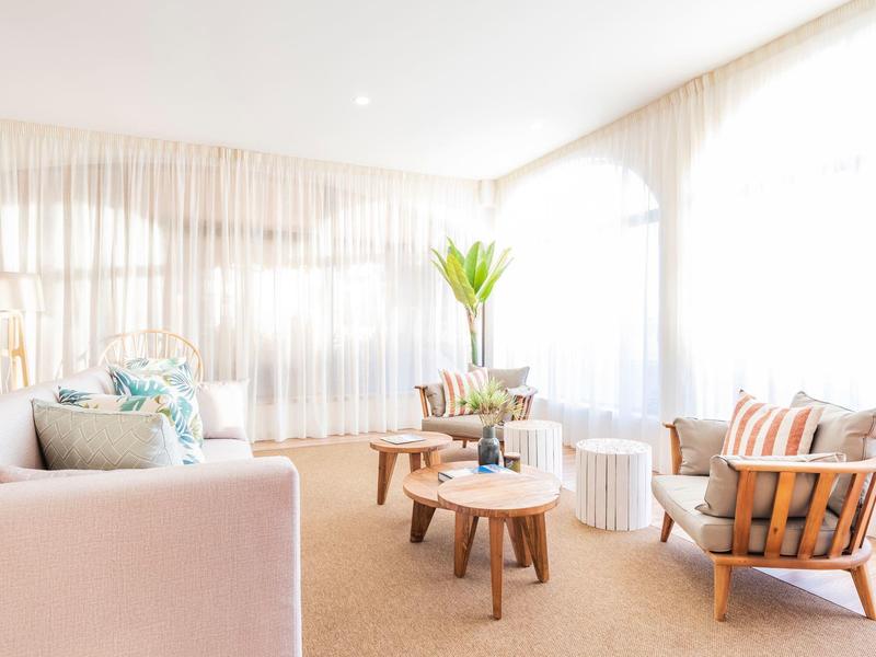 Jasny salon z wygodnymi fotelami, drewnianymi stolikami i dużymi oknami z zasłonami.