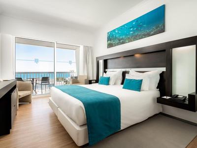 Moderne hotelkamer met groot bed, balkon, uitzicht op zee en blauwe accenten.