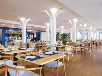 Restaurante de hotel moderno y luminoso con mesas de madera, sillas blancas, grandes ventanas y suelo de madera.