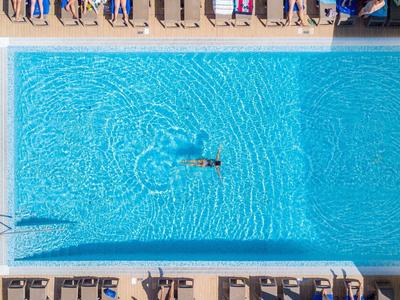 Piscina cuadrada con agua clara y una persona nadando, rodeada de tumbonas.