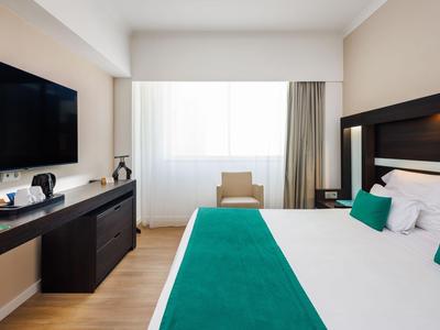Habitación de hotel moderna con cama grande, corredor verde, televisor y ventana con cortinas.