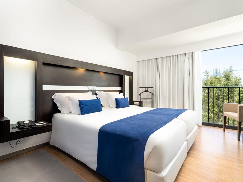 Lichte hotelkamer met een groot bed, blauwe accenten en balkonzicht op groen.