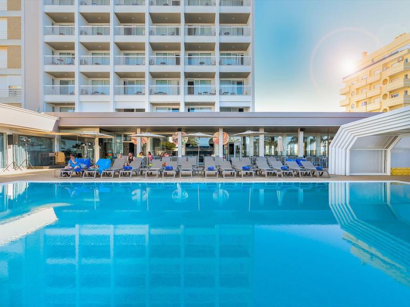 Hotelzwembad met ligstoelen en moderne gebouwen onder een heldere lucht.