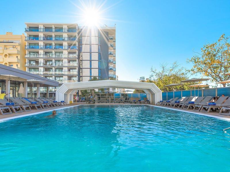 Zonnig buitenzwembad met ligstoelen en modern hotelgebouw op de achtergrond.