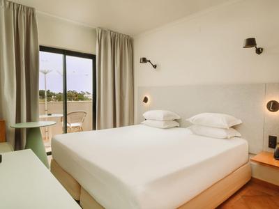 Hell eingerichtetes Hotelzimmer mit großem Bett, Nachttischen und Blick auf die Terrasse.