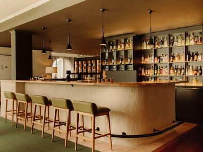Moderner Hotelbarbereich mit Holztheke und fünf Hockern mit grünen Sitzen.