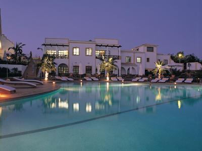 Beleuchteter Hotelpool mit Liegestühlen und weißen Gebäuden bei Dämmerung.