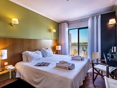 Bright hotel room with double bed, bedside lamps, balcony, and outdoor view of trees and buildings.