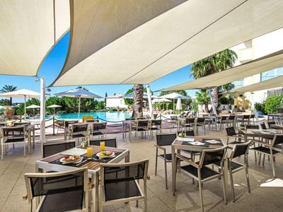Outdoor hotel dining area with modern tables, chairs, shade sails, and pool view.