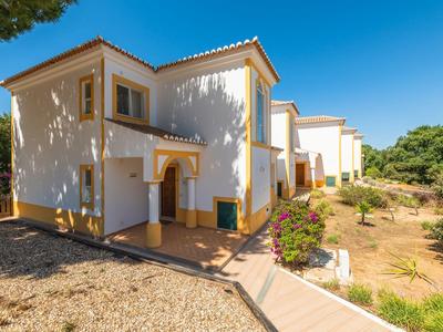 White two-story house with tiled roof under a clear blue sky, surrounded by dry landscape and greenery.