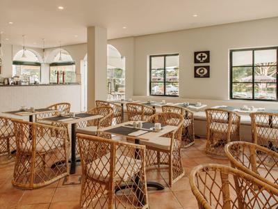 Helles Hotelrestaurant mit Korbstühlen und Tischen, großen Fenstern und Blick auf Berge.