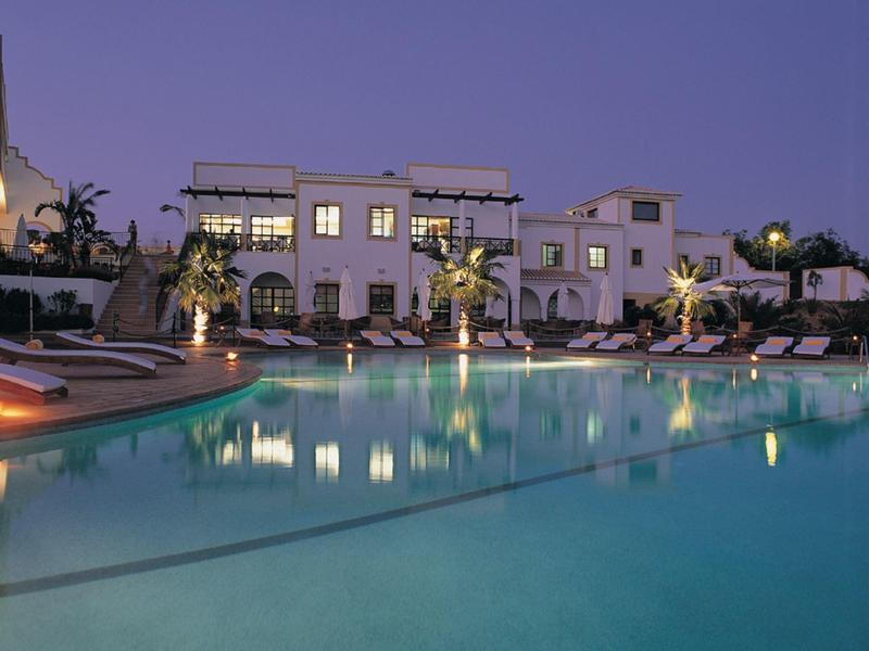 Hotel pool at dusk with lit palm trees and white lounge chairs reflecting in water.