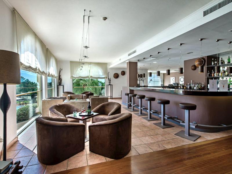 Cozy hotel bar with leather chairs, round tables, large windows, and modern lighting.