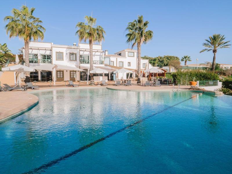 Resort with clear blue pool, palm trees, and white buildings under a blue sky.