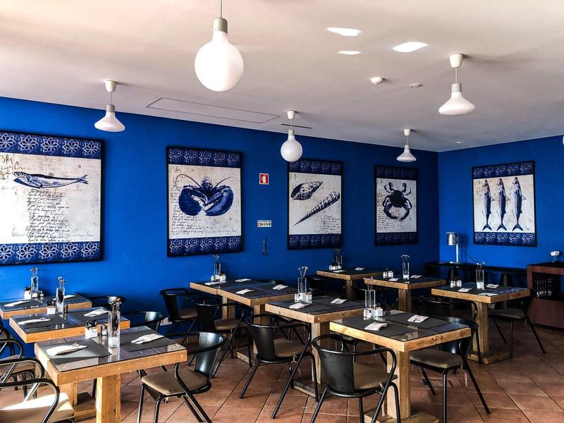 Dining area with wooden tables, black chairs, blue walls, and seafood-themed artwork under white pendant lights.