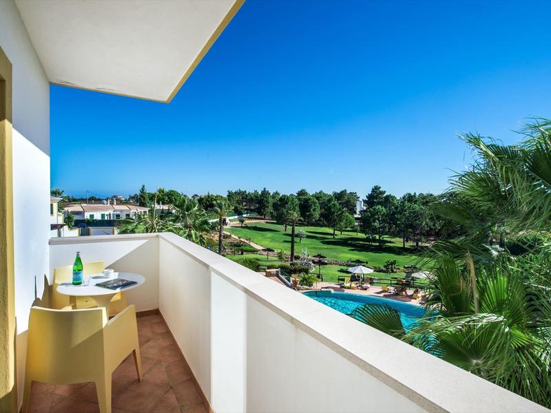 Balcony with table and chairs overlooking a green golf course and blue pool under clear sky.