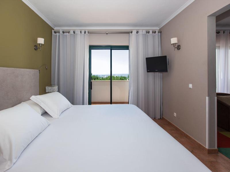 Modern hotel room with large bed, balcony door, wall-mounted TV, and bedside lamps.