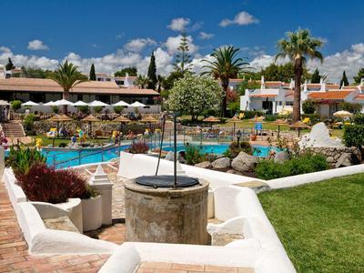 Complesso alberghiero con piscina, ombrelloni, palme e prati verdi sotto un cielo blu.