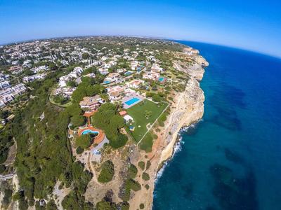 Vista aerea di una zona costiera con ville, piscine e mare azzurro limpido in una giornata di sole.
