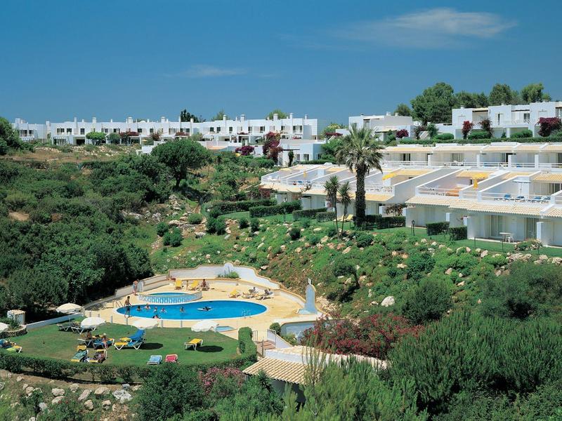 Complesso alberghiero con giardino, piscina e edifici bianchi sotto un cielo azzurro.