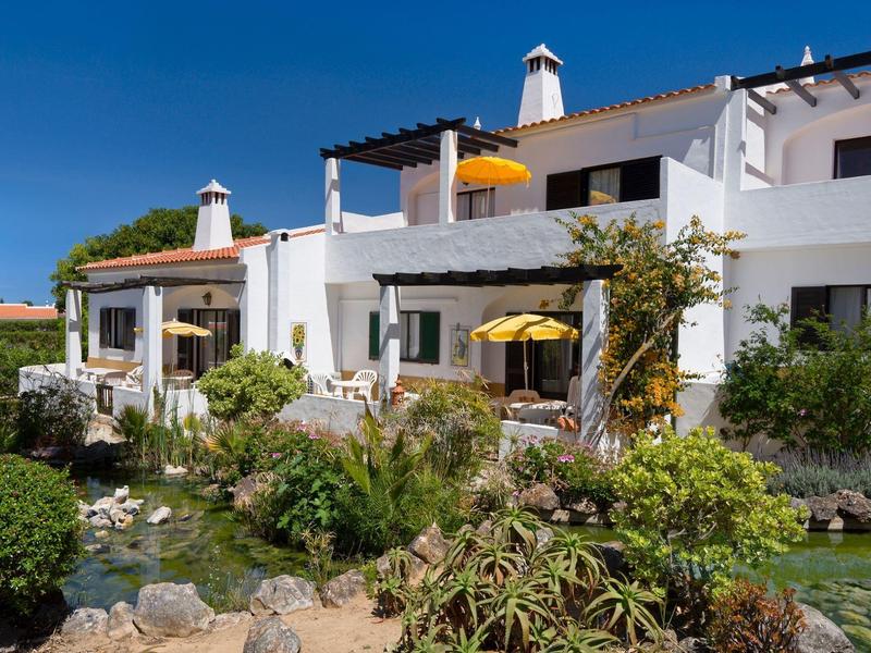 Casa vacanze bianca con accenti gialli solari e giardino rigoglioso sotto il cielo blu.